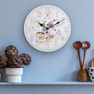 Lifestyle image of the 404-2631T 12-inch Teatime Wall Clock shown hanging on a sky blue wall above a shelf holding a plant and wooden spoons. The clock features an image of a floral teapot and teacup on a light-textured background, with the text "Tea Time," white numbers, black metal hands, and an open face design.