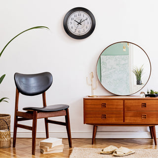 Lifestyle image of the 404-2636 14-inch Savannah Wall Clock shown hanging on a white wall in a mid-century modern decorated room with a black leather chair and dresser with a round mirror. The clock features a parchment-inspired dial with traditionally classic black numbers, straight black metal hands, and an oil-bronze decorative frame.