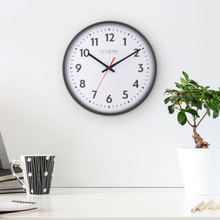Lifestyle image of the 404-2636 13-inch Commercial Wall Clock shown hanging on a white wall above a white modern desk with a balck coffee cup and a plant. The clock features bold black numerals, black hour markings, a white dial, black hour and minute hands, and a red second hand in a black flush-mount frame.