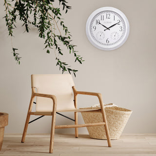 Lifestyle image of the 404-3015CC 15-inch Indoor/Outdoor Payton Wall Clock shown hanging on a beige wall above a light wood armchair with a cream seat. A woven basket sits on the floor nearby, with green hanging leaves entering from the top left. The clock features a white distressed frame, a white dial with black Arabic numerals, black hands, and two small sub-dials for temperature and humidity.