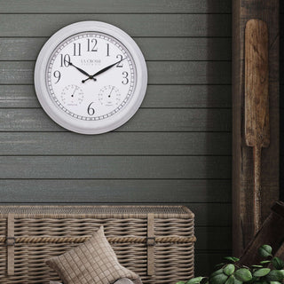 Lifestyle image of the 404-3015CC 15-inch Indoor/Outdoor Payton Wall Clock shown on a dark gray wood-paneled wall above a woven rattan bench with a beige pillow. The clock features a white distressed frame, a white dial with black Arabic numerals, black hands, and two small sub-dials for temperature and humidity.
