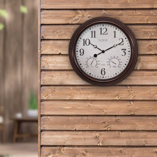 Lifestyle image of the 404-3015 15-inch Indoor Outdoor Wall Clock shown hanging on a rustic wood plank building with a patio in the background. The clock features a linen-inspired dial with stylized numbers and hands, with temperature and humidity dials surrounded by a dark brown finish frame.