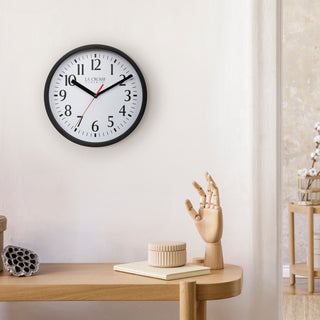 Lifestyle image of the 404-3020B 8-inch Basics Wall Clock - Black shown hanging on a light wall above a wood side table with a wood artist's hand and a book. The clock features a black flush-mount frame, a white dial with black numbers, black hour and minute hands, and a red second hand.