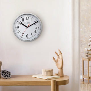 Lifestyle image of the 404-3020S 8-inch Basics Analog Wall Clock – Silver shown hanging on a light wall above a wood side table with a wood artist's hand and a book. The clock features a black flush-mount frame, a white dial with black numbers, black hour and minute hands, and a red second hand.