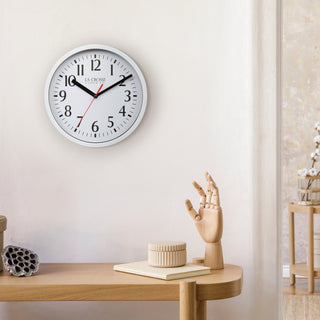 Lifestyle image of the 404-3020W 8-inch Basics Analog Wall Clock – White shown hanging on a light wall above a wood side table with a wood artist's hand and a book. The clock features a white flush-mount frame, a white dial with black numbers, black hour and minute hands, and a red second hand.