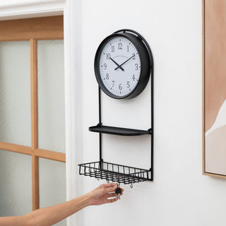 Lifestyle image of the 404-3022 Depot Wall Clock with Basket and Shelf hanging in an entryway showing someone hanging keys on the "S" hook. The clock features a Black Metal Frame with a shelf and a basket, an archival paper-inspired dial, black numbers, and black metal hour and minute hands.