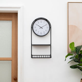 Lifestyle image of the 404-3022 Depot Wall Clock with Basket and Shelf hanging in an entryway with a plant and picture. The clock features a Black Metal Frame with a shelf and a basket, an archival paper-inspired dial, black numbers, and black metal hour and minute hands.