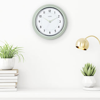 Lifestyle image of the 404-3024PH 9.5-inch Diner Wall Clock - Pistachio shown hanging on a white wall above a white shelf with books and plants. The clock features an off-white dial with black numbers, pistachio hour dots, pistachio hour and minute hands, and a silver accent second hand set within a vintage-retro pistachio green frame.