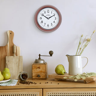 Lifestyle image of the 404-3024TC 9.5-inch Diner Clock – Terracotta shown hanging on a light grey wall above a wood side table with an antique coffee grinder, a white ceramic pitcher, pears, coffee, and other kitchen items. The clock features a retro-style terracotta frame with a white dial, chrome bezel, black numbers and minute markings, black metal hands, and a silver second hand.
