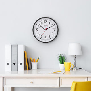Lifestyle image of the 404-3025B 10-inch Basic Decor Wall Clock with a Black frame, white dial with black numbers, hour, and minute hands, and a red second hand hanging on a white wall above a white desk with a lamp, notebook, pencil, desk accessories, a yellow coffee mug, and a yellow chair