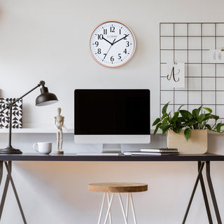 Lifestyle image of the 404-3025C 10-inch Basic Wall Clock – Copper, hanging on a light tan wall above a black desk. The desk has a computer, notebooks, a plant, a coffee mug, a black desk lamp, and a posable wood model. The clock features a white dial, black numbers, markings, hour, minute, and second hands surrounded by a copper color flush-mount frame.