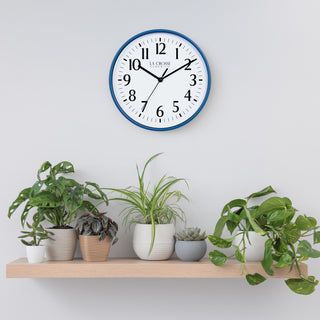 Lifestyle image of the 404-3025NB 10-inch Basic Wall Clock - Navy Blue hanging on a light grey wall above a pine shelf with multiple plants. The clock features a white dial, black numbers and markings, and hour, minute, and second hands, surrounded by a navy blue flush-mount frame.