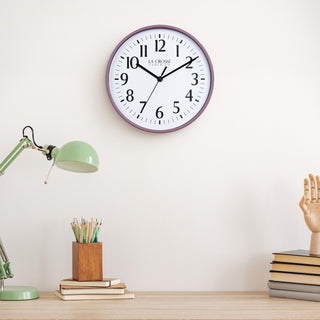 Lifestyle image of the 404-3025RR 10-inch Basic Wall Clock – Ruby Red, hanging on a light tan wall above a light wood desk with a green desk lamp, a wood pencil holder on top of a stack of notebooks, and a wood modeling hand sitting on top of a pile of books. The clock features a white dial, black numbers, markings, hour, minute, and second hands surrounded by a ruby red rose flush-mount frame.