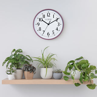 Lifestyle image of the 404-3025RR 10-inch Basic Wall Clock - Ruby Red hanging on a light grey wall above a pine shelf with multiple plants. The clock features a white dial, black numbers and markings, and hour, minute, and second hands, surrounded by a ruby red rose flush-mount frame.