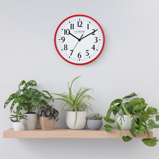 Lifestyle image of the 404-3025R 10-inch Basic Wall Clock - Red hanging on a light grey wall above a pine shelf with multiple plants. The clock features a white dial, black numbers and markings, and hour, minute, and second hands, surrounded by a red flush-mount frame.