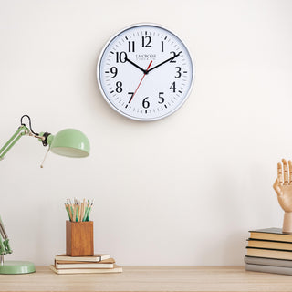 Lifestyle image of the 404-3025S 10-inch Basic Wall Clock – Silver, hanging on a light tan wall above a light wood desk with a green desk lamp, a wood pencil holder on top of a stack of notebooks, and a wood modeling hand sitting on top of a pile of books. The clock features a white dial, black numbers, markings, hour, and minute hands, and a red second hand, surrounded by a silver flush-mount frame.