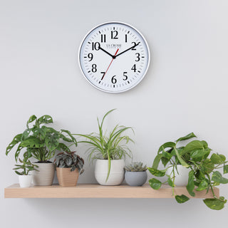 Lifestyle image of the 404-3025S 10-inch Basic Wall Clock - Silver hanging on a light grey wall above a pine shelf with multiple plants. The clock features a white dial, black numbers and markings, hour, and minute hands, and a red second hand, surrounded by a silver flush-mount frame.