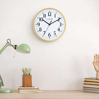 Lifestyle image of the 404-3025Y 10-inch Basic Wall Clock – Yellow, hanging on a light tan wall above a light wood desk with a green desk lamp, a wood pencil holder on top of a stack of notebooks, and a wood modeling hand sitting on top of a pile of books. The clock features a white dial, black numbers, markings, hour, minute, and second hands surrounded by a yellow flush-mount frame.
