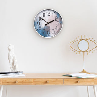 Brushstroke 404-3028A 11 inch wall clock featuring a white case surrounding a painted brush design dial in blues creams and taupe black numbers and black metal hands hanging on a wall above a side table desk and a white statue and mirror eyeball decoration