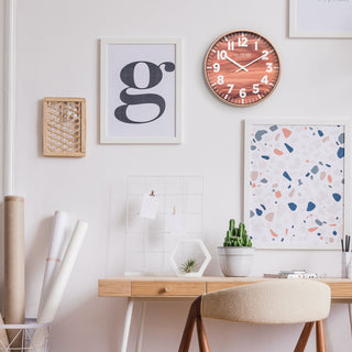 Lifestyle image of the 10.8-inch Atwood Wall Clock hanging on a wall in a home office setting with a neutral desk, fun wall art, and desk accessories. The clock features a copper finish frame, a wood design dial, white numbers, and white metal hour and minute hands.