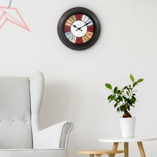 Lifestyle image of the 404-3030A 12-inch Gatherings Wall Clock shown hanging on a wall above a chair and side table. The clock features a black decorative frame, a multi-color segmented dial with Roman numerals, and black hour and minute hands.