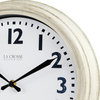 Close-up image of the frame of the 404-3030W 12-inch Wynn Wall Clock. The image shows the detail of the off-white weathered frame surrounding a white dial with distressed black numbers and decorative black metal hands with the La Crosse Clock logo.