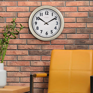 Lifestyle image of the 404-3030W 12-inch Wynn Wall Clock shown hanging on multi-colored brick wall above a goldenrod leather chair next to a side table with a plant. The clock features an off-white weathered frame surrounding a white dial with distressed black numbers and decorative black metal hands with the La Crosse Clock logo.