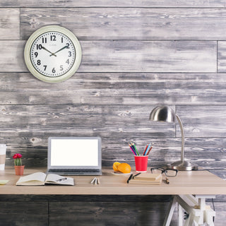 Lifestyle image of the 404-3030W 12-inch Wynn Wall Clock shown hanging on a grey barnboard panelled wall above a light colored neutral desk with a laptop and lap. The clock features an off-white weathered frame surrounding a white dial with distressed black numbers and decorative black metal hands with the La Crosse Clock logo.