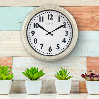 Lifestyle image of the 404-3030W 12-inch Wynn Wall Clock shown hanging on a multi-colored panelled wall above a cream countertop with white potted succelant plants. The clock features an off-white weathered frame surrounding a white dial with distressed black numbers and decorative black metal hands with the La Crosse Clock logo.