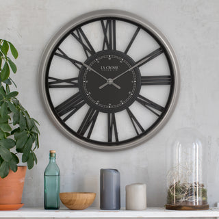 A large round wall clock with a brushed silver frame, black Roman numerals, and a black center dial with silver hands. The clock is mounted on a light gray textured wall above a shelf with decorative items including a potted plant, glass bottle, bowl, candles, and a glass dome.