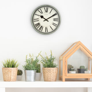 A round wall clock with a gray frame and Roman numerals hangs on a white wall above a shelf with potted plants and a small wooden house planter.