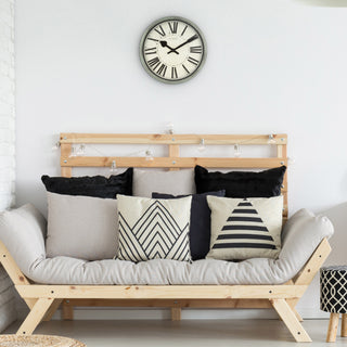 Lifestyle image of the 404-3036G 14-inch Sagebrook Wall Clock shown hanging on a white wall above a natural wood loveseat with neutral and black geometric pillows. The clock features a sage green distressed decorative frame, an ivory linen design dial, bold black Roman numerals, and decorative black metal hour and minute hands.