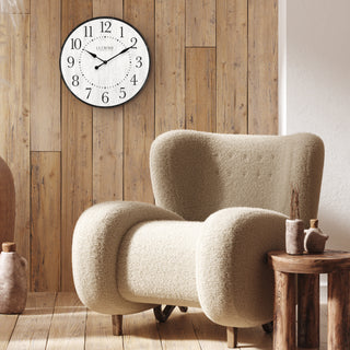 A modern beige bouclé armchair in a rustic living room setting with a wooden plank wall, a round white wall clock, and a small wooden side table.