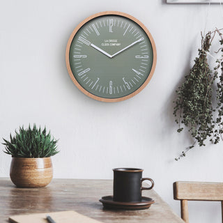 Lifestyle image of the 404-3041D 16-inch Ellwood Wall Clock, with Silent Sweep, shown hanging on a wall aboe a dining table with a chair, the table has plants, and a coffee cup. The clock features a flush-mount light wood frame, a sage green dial, modern white numbers, and white metal hour and minute hands.