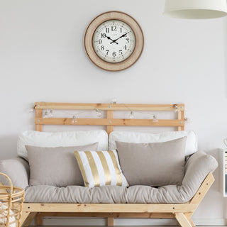 A light natural wood daybed with a slatted backrest, styled with beige and white pillows, including a striped accent pillow. A round wooden wall clock hangs above on a white wall.