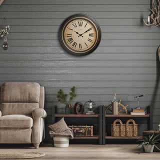 Rustic living room with dark horizontal paneled walls with a side chair and the 404-3045BK 18-inch Traditions Wall clock featuring a dark decorative frame with bronze bezel surrounding a linen detailed dial with black Roman numerals and metal decorative hands hanging on the wall above low bookshelves