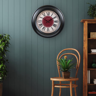 Lifestyle image of the 404-3045B 18-inch Barton Wall Clock, hanging on an evergreen slatted wall with a wooden country-style dining chair and bookshelf. The clock features a parchment-inspired dial with a burgundy-red center, surrounded by black Roman numerals, within a decorative dark espresso-brown frame.