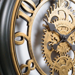 Close-up of a decorative wall clock showing ornate gold-toned numerals and interlocking gear cogs with dark gray hands against a light background.