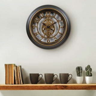 A round wall clock with a dark gray frame and ornate gold-toned gears in the center, mounted on a light gray wall above a wooden shelf with books, mugs, and small cactus plants.
