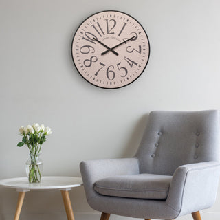 404-3053 A round wall clock with a pale pink face and black numerals and hands, mounted on a light gray wall above a gray upholstered armchair with a small white side table holding a vase of white flowers.