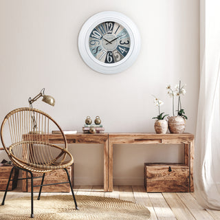 404-3056 A round wall clock with a white frame and distressed blue-gray numerals hangs above a rustic wooden console table in a styled room with a wicker chair, lamp, and potted orchids.