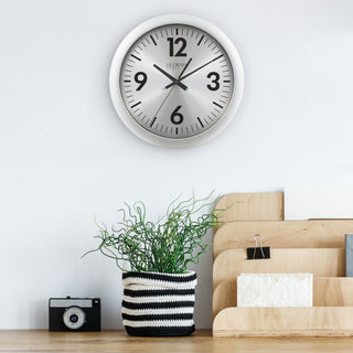 404-3229 A round wall clock with a silver-toned frame and a white face, mounted on a white wall above a wooden surface with a potted green plant, a small camera, and light wood organizers.