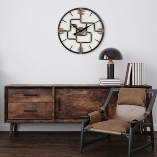 Lifestyle image of the 404-3420 20-inch Pipeworks Metal Wall Clock shown hanging on a white wall above a Mid-Century Modern console table with a retro black lamp and a brown architectural chair. The clock features an open face dial with a weathered bronze and antique brass finish pipe framework, and antique brass numbers 3, 6, 9, and 12.