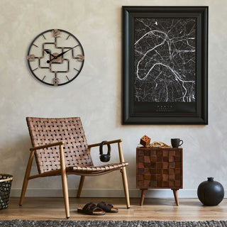 Lifestyle image of the 404-3420 20-inch Pipeworks Metal Wall Clock shown hanging on a tan plaster finish wall hanging next to map artwork, and above a dark wicker chair with a woven wood side table. The clock features an open face dial with a weathered bronze and antique brass finish pipe framework, and antique brass numbers 3, 6, 9, and 12.