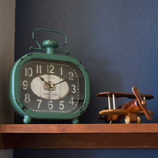 404-3425 A distressed vintage-style teal metal alarm clock sits on a wooden shelf against a dark blue wall, next to a small wooden biplane toy.