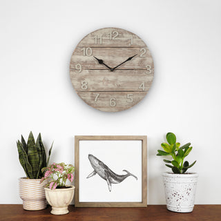 404-3430W A round rustic wood wall clock with black hands and numbers hangs on a white wall above a wooden surface with potted plants and a framed whale illustration.