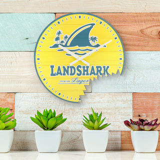 Lifestyle image of the 404-3435 14-inch Margaritaville Landshark MDF Wall Clock shown hanging on a multi-colored weathered wood paneled wall above a white quartz counter with potted plants. The clock features an open-face faux yellow weathered wood dial with a "bite" taken out of it, the Landshark Premium Lager logo, and white hands with blue time markings.