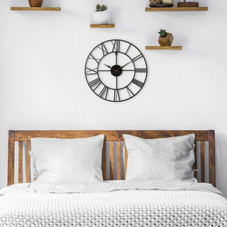 Lifestyle image of the 404-3451 20-inch Metal Tower Wall Clock - Roman Numerals, shown hanging on a white wall above a wooden slatted bed with white bedding. The clock features an open-face cutout design, Roman numerals, metal hands, and a metal skeleton frame.