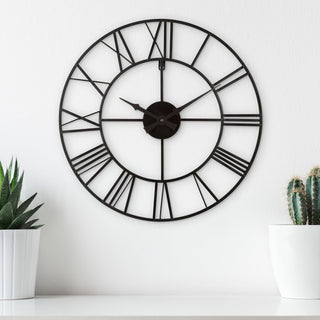 404-3451 A large black metal wall clock with Roman numerals hangs on a white wall above a white shelf, flanked by potted plants on both sides.