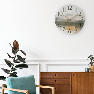 Lifestyle image of the 404-3461 24-inch Daybreak Wall Clock shown in a mid-century modern living room on a white wall above a teal blue chair and mid-tone wood sideboard. The clock features an open face design of an impressionistic sunrise in muted tones, distressed numbers, and gold metal hands.
